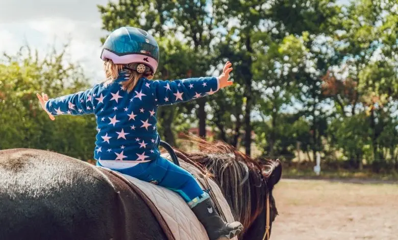 Dívka v helmetě jezdí na koni venku s rozpaženýma rukama.