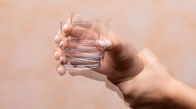 Ruka drží sklenici vody na neutrálním pozadí.
