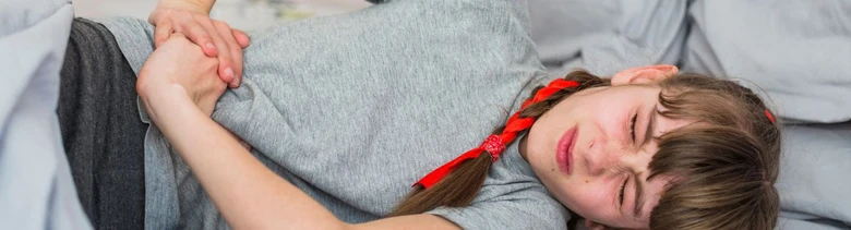 Dívka leží v bolesti na posteli s rukama na boku.