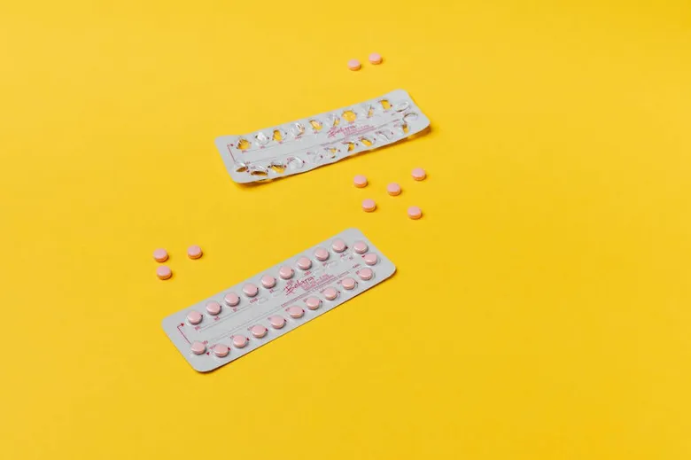 Dvě balení hormonální antikoncepce s tabletami na žlutém pozadí.