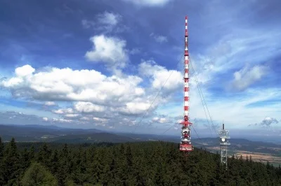 Vysílač na kopci v lesu pod modrou oblohou s mraky.