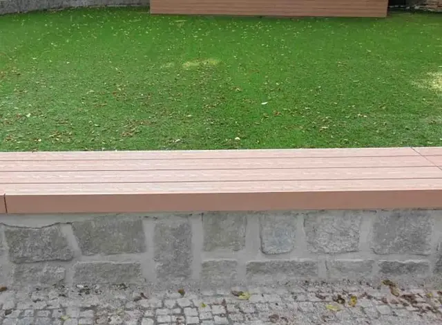 Lavička z dřevoplastu na kamenném podkladě v zahradním prostředí.