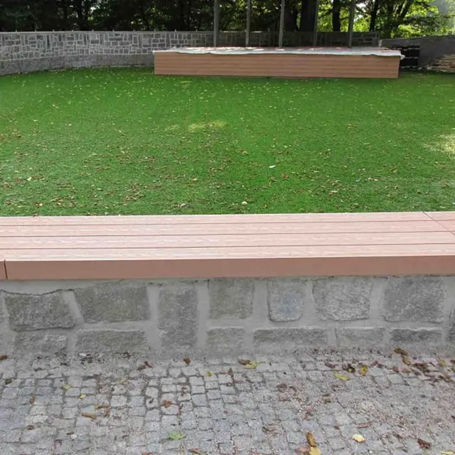 Lavička z dřevoplastu na kamenném podkladě v zahradním prostředí.