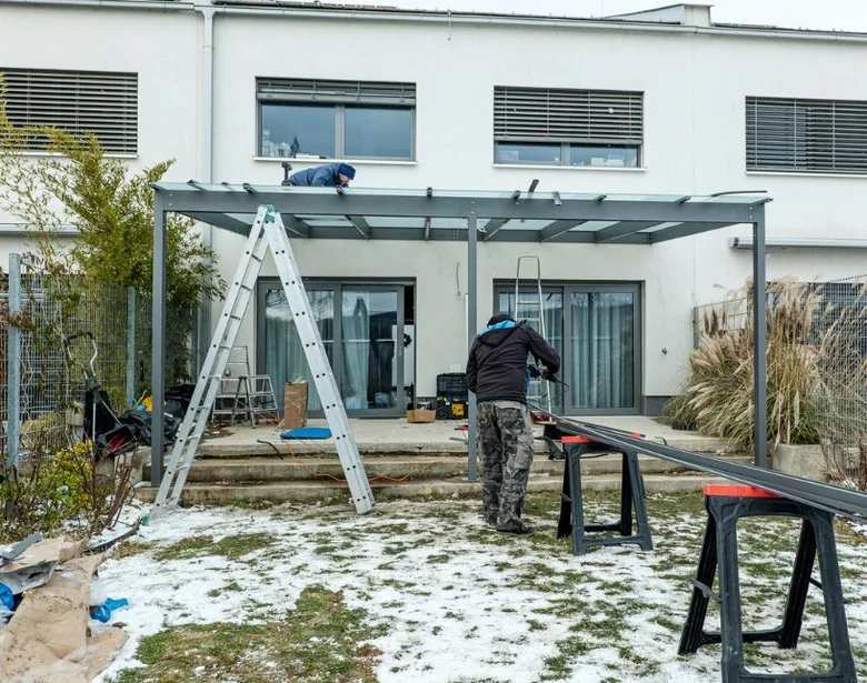 Dva dělníci montují kovovou konstrukci pergoly v zimním zahradě.